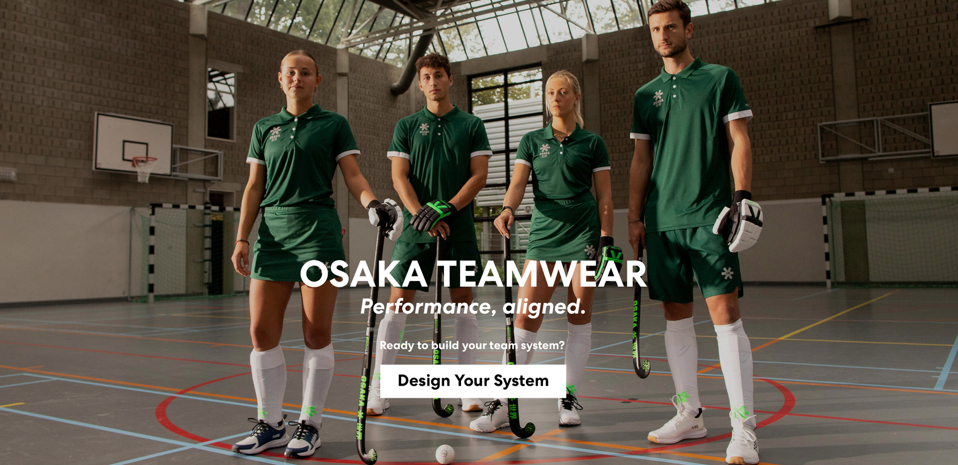 Four field hockey players wearing matching Osaka custom green team kit — polo shirts, shorts, and skort — holding hockey sticks and gloves inside an indoor sports hall