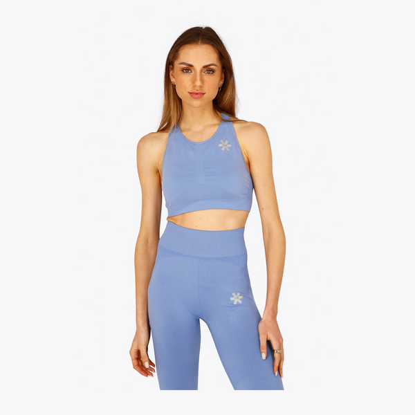 A woman modeling a light blue athletic set consisting of a halter crop top and high-waisted leggings, both featuring a small white flower logo on the chest and hip, against a plain light gray background. #ManorBlue