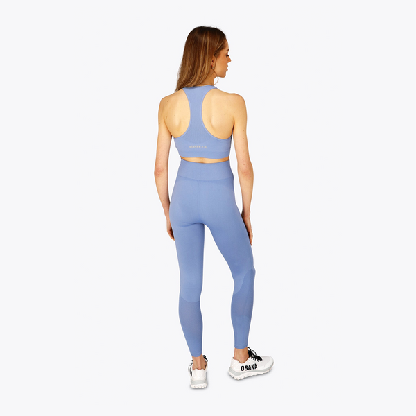 A woman is shown from the back wearing a matching light blue athletic set consisting of a sports bra and high-waisted leggings, paired with white sneakers, against a plain light gray background. #ManorBlue