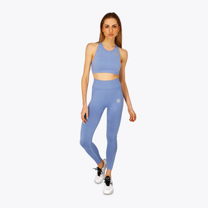 A woman modeling a matching light blue activewear set consisting of a sports bra and high-waisted leggings, with a small white logo on the chest and hip, set against a plain light gray background. #ManorBlue