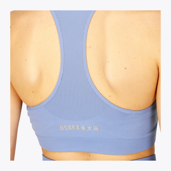 Back view of a person wearing a light blue athletic sports bra with the brand name OSAKA printed in white near the lower back. #ManorBlue