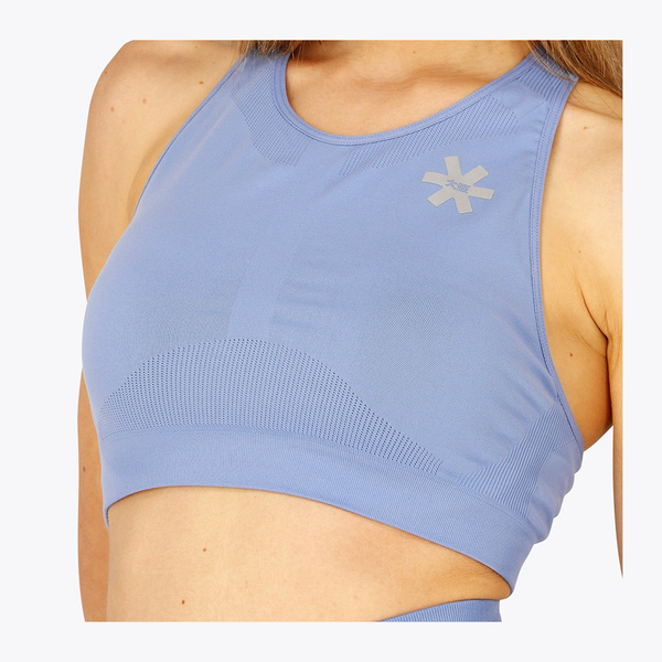 A close-up of a woman wearing a light blue athletic sports bra with a textured knit pattern and a small reflective logo on the chest. #ManorBlue