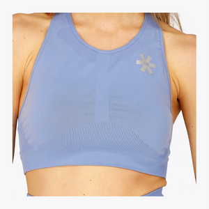 A close-up of a woman wearing a light blue athletic sports bra with a small silver logo on the upper chest, set against a plain white background. #ManorBlue