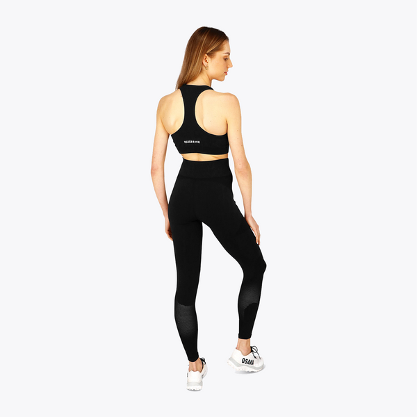 A woman is shown from the back wearing a black sports bra and matching high-waisted leggings with black textured panels on the lower legs, paired with white sneakers, against a light gray background. #FadedBlack