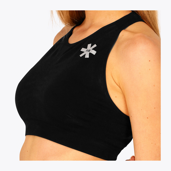 Close-up of a woman wearing a black ribbed sports bra with a small silver geometric logo on the upper chest, against a plain light background. #FadedBlack