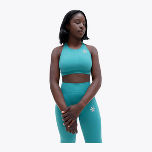 A woman modeling a matching teal athletic set consisting of a halter crop top and high-waisted leggings, both featuring a small white flower logo, against a solid white background. #BayouGreen