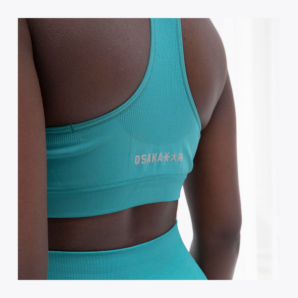 Close-up back view of a person wearing a teal sports bra and matching high-waisted bottoms, with the brand name OSAKA printed in white on the sports bra. #BayouGreen
