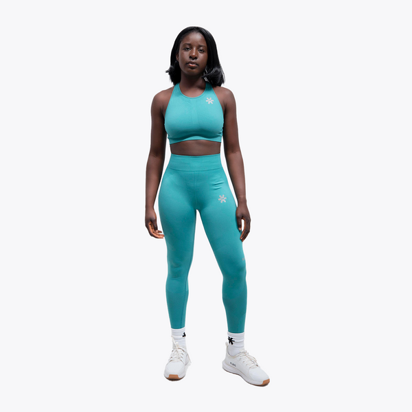 A woman modeling a matching turquoise athletic set consisting of a halter crop top and high-waisted leggings, with small white snowflake graphics on the chest and upper thigh. She is wearing white socks and white sneakers against a solid light gray background. #BayouGreen