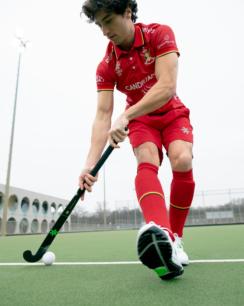 Male field hockey player in Osaka custom red national team kit — polo shirt, shorts, and socks with gold trim — dribbling with an Osaka hockey stick on an outdoor turf pitch