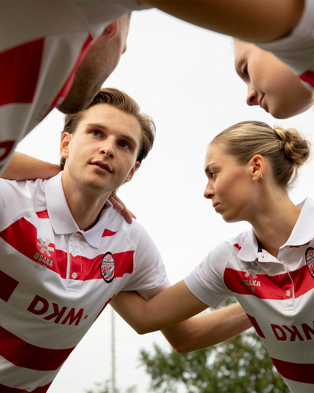 Sports team huddle wearing Osaka custom white and red striped club kit with sponsor branding, showing fully customizable teamwear with club badge and logo placement