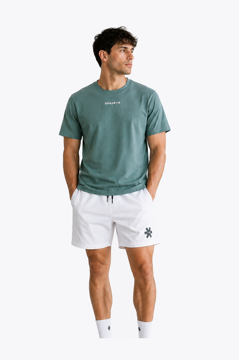 Male model wearing Osaka Men’s Apex Training Tee in Pineneedle green with white training shorts, showcasing reflective chest logo and breathable athletic performance fabric.
