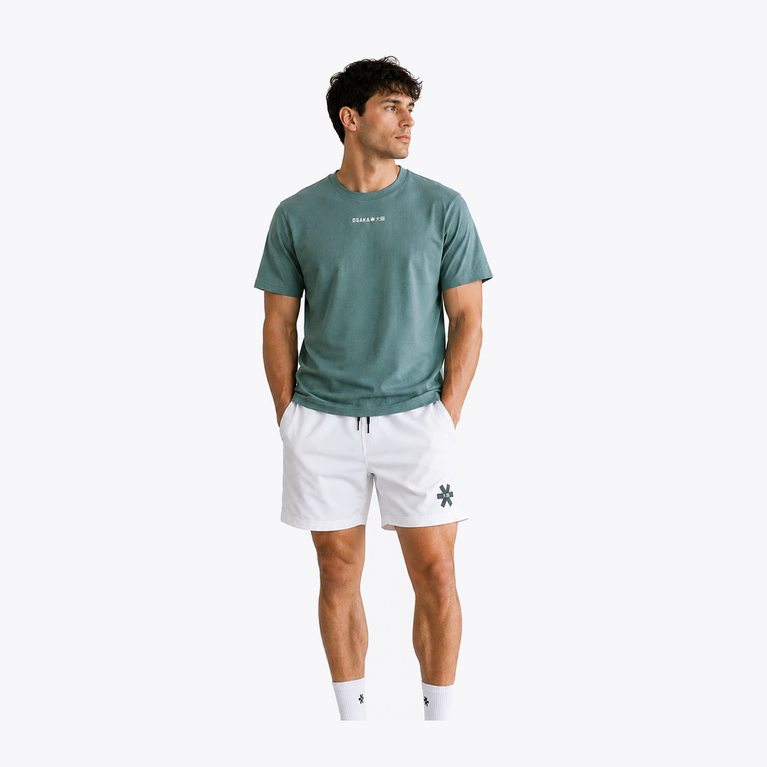 Male model wearing Osaka Men’s Apex Training Tee in Pineneedle green with white training shorts, showcasing reflective chest logo and breathable athletic performance fabric.