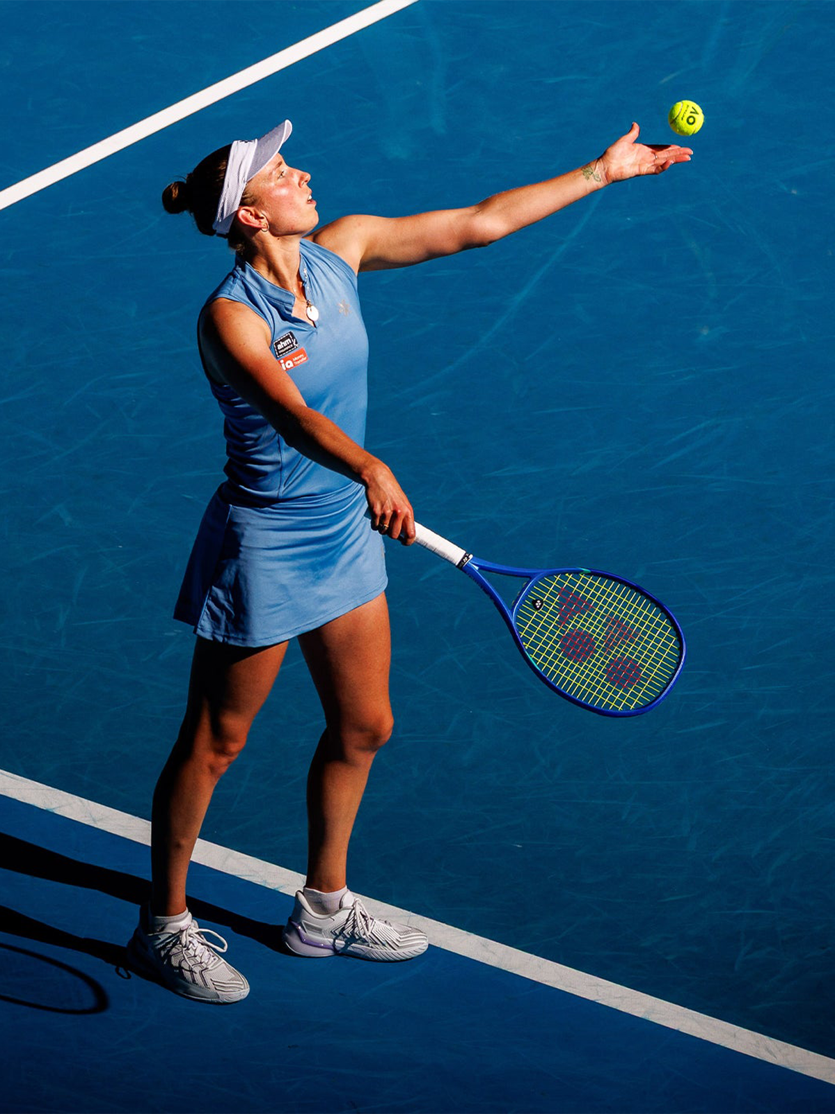 Elise Mertens serving on a blue hard court, tossing the tennis ball overhead with racket ready