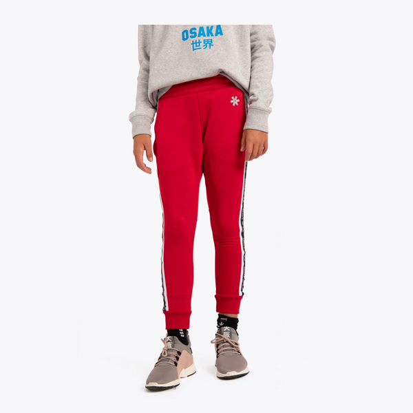 A model wearing red athletic jogger pants with white and black side stripes, paired with a gray sweatshirt and beige sneakers, shown from the waist down on a light gray background.