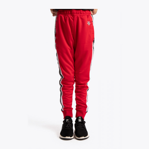 Red sweatpants with black and white side stripes and a small white flower logo on the upper left thigh, shown on a model from the waist down wearing black sneakers against a light gray background.