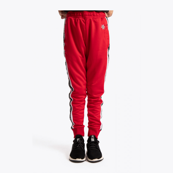 Red sweatpants with black and white side stripes and a small white flower logo on the upper left thigh, shown on a model from the waist down wearing black sneakers against a light gray background.