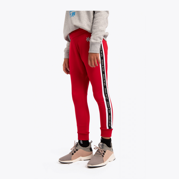 A child model wearing bright red athletic jogger pants with a white and black side stripe, paired with a gray sweatshirt and beige sneakers, shown against a plain light gray studio background.