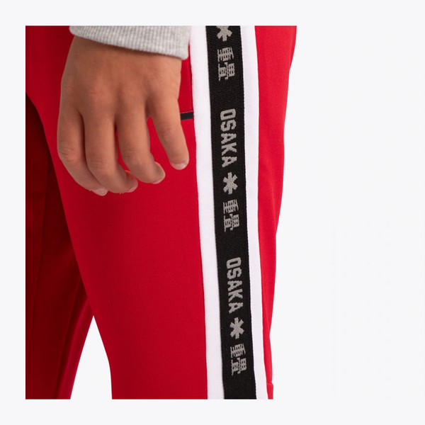 Close-up of a person wearing bright red track pants with a black and white DSAKA brand stripe down the side, showing a hand and part of a light gray top.