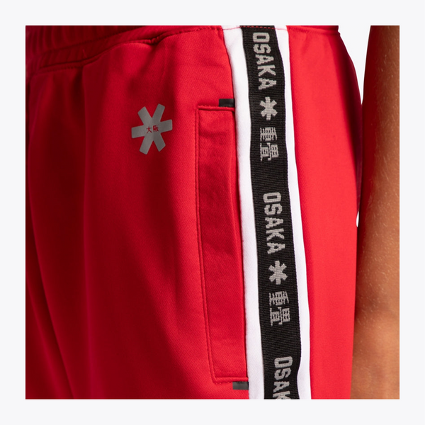 Close-up of red athletic shorts with a black and white side stripe featuring repeated white 'OSAKA' text and a gray logo, shown on a model.