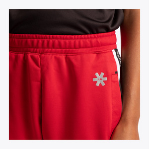 Close-up of bright red athletic shorts with a silver reflective star logo on the left leg, worn with a black top. A black side tag with white text is visible on the right side.
