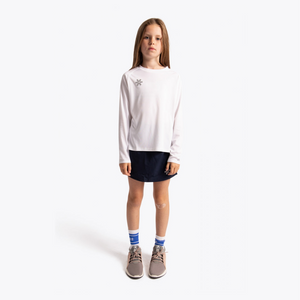 A young girl stands against a light gray studio background wearing a white long-sleeve top with a small silver graphic on the chest, a dark navy skirt, blue and white striped socks, and gray sneakers. #White