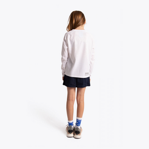 A child model shown from behind wearing a white long-sleeve athletic top, a dark navy athletic skirt, white socks with blue stripes, and sneakers, against a clean white studio background. #White