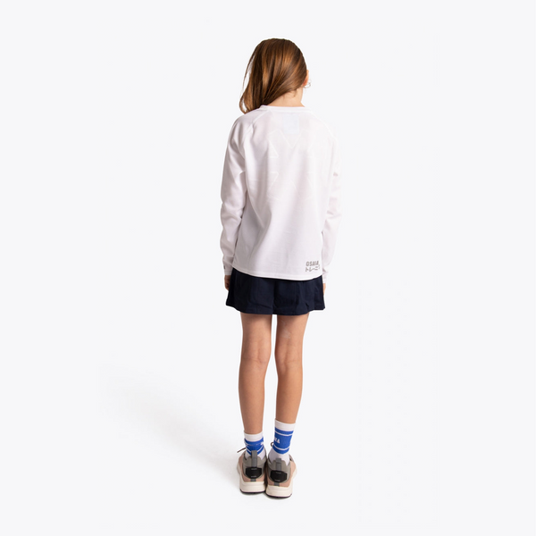 A child model shown from behind wearing a white long-sleeve athletic top, a dark navy athletic skirt, white socks with blue stripes, and sneakers, against a clean white studio background. #White