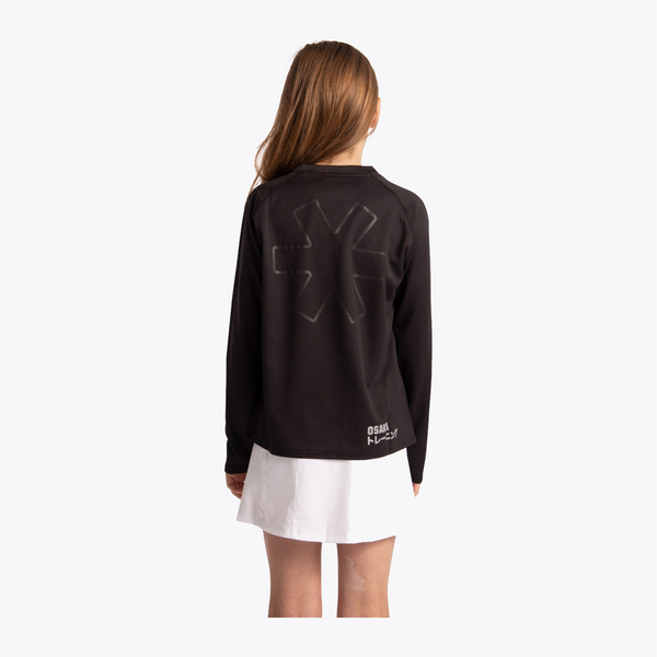 A girl viewed from the back wearing a black long-sleeve shirt with a large tonal black graphic on the back and a white skirt, against a plain light gray studio background. #Black

