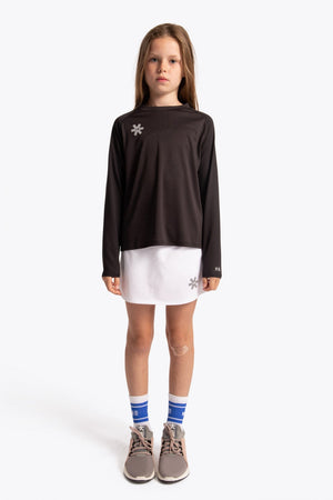 A young girl stands against a light gray studio background wearing a dark brown long-sleeve athletic top with a small white snowflake logo on the left chest, a white mini skirt, blue and white striped socks, and gray sneakers. #Black