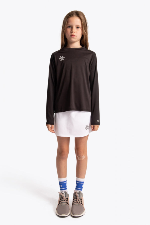 A young girl stands against a light gray studio background wearing a dark brown long-sleeve athletic top with a small white snowflake logo on the left chest, a white mini skirt, blue and white striped socks, and gray sneakers. #Black