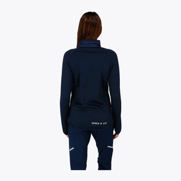 A woman viewed from the back wearing a dark navy athletic jacket and matching navy pants against a light gray studio background. #Navy