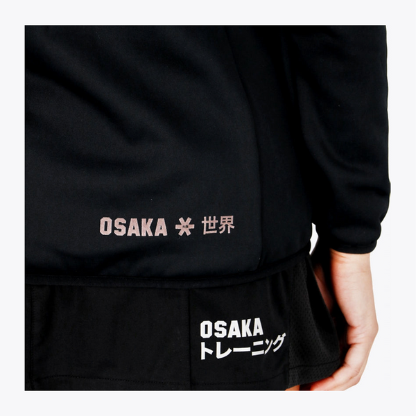 Close-up of a black long-sleeve athletic top or training jacket with printed text. The upper chest area shows 'OSAKA' in light pink with Japanese characters, and the lower hem has a black mesh pocket with 'OSAKA' and Japanese text in white. #Black
