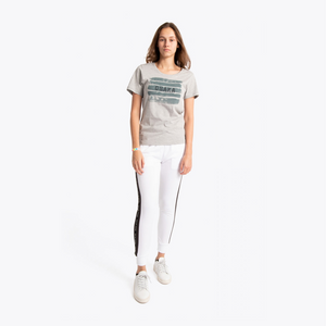 A woman modeling a light gray graphic t-shirt with horizontal stripes and the word 'STUDIOS' printed on the chest, paired with white pants featuring black side stripes and white sneakers, set against a plain white studio background. #White