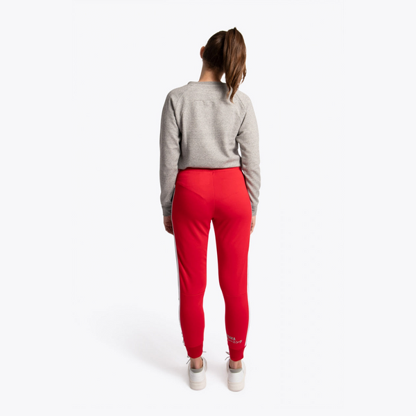 A woman is shown from behind wearing a gray cropped sweatshirt and bright red athletic jogger pants with white stripes down the sides, standing against a plain white studio background. #Red