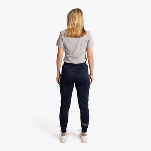 A woman viewed from behind wearing a light gray t-shirt, navy blue pants, and white sneakers against a plain white studio background. #Navy