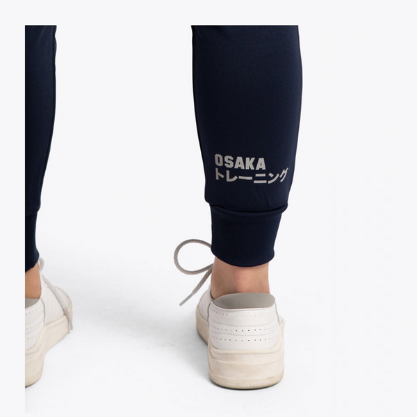 Close-up back view of a person wearing dark navy jogger pants with white 'OSAKA トレーニング' text on the lower calf, paired with off-white slip-on sneakers against a clean white background. #Navy