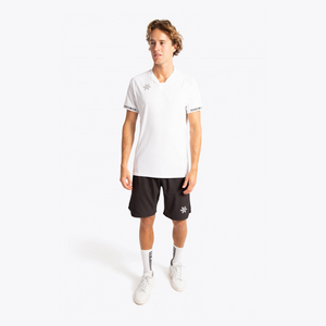 A male model stands against a light gray studio background wearing a white short-sleeve athletic polo shirt with a small star logo on the chest and sleeve trim, black athletic shorts with a small star logo, white socks, and white sneakers. #White