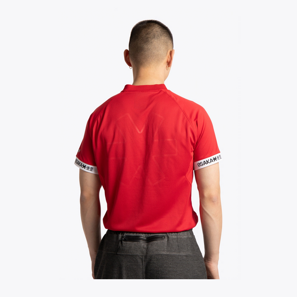 A man viewed from behind wearing a bright red short-sleeve athletic shirt with white trim on the sleeves and dark gray pants, against a plain light gray studio background. #Red