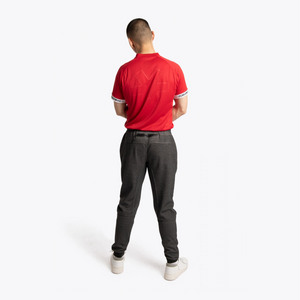 A man viewed from behind wearing a bright red short-sleeve polo shirt with white trim on the sleeve cuffs, dark gray slim-fit pants, and white sneakers, against a plain white studio background. #Red