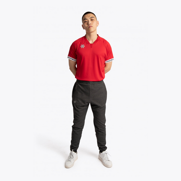 A model stands against a light gray studio background wearing a red short-sleeve athletic polo shirt with white trim and a small logo on the chest, paired with dark gray jogger pants and white sneakers. #Red