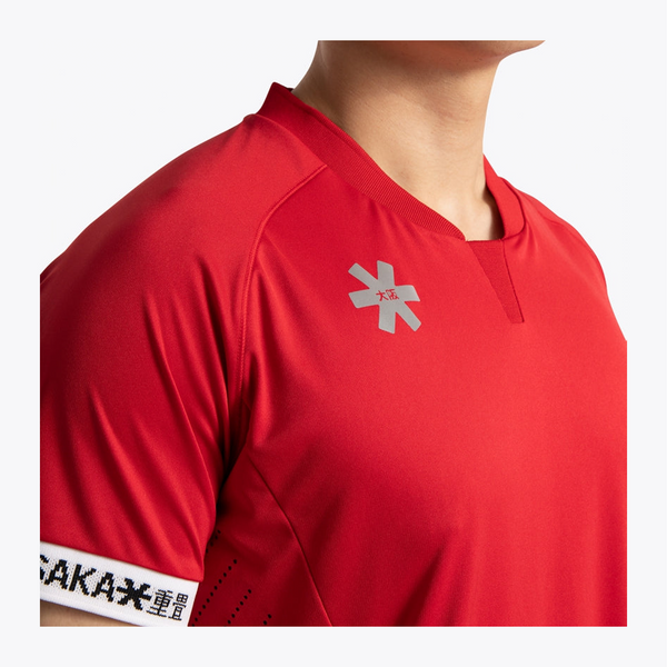 Close-up of a red athletic jersey worn by a model, showing the chest with a light gray logo and part of the sleeve with black text. #Red